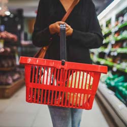 Close-up photo of a woman's hands carrying a red basket down the produce aisle at a grocery store.