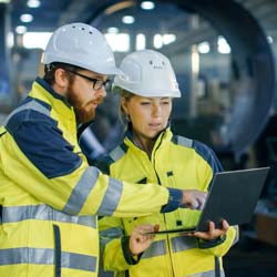 A man and a woman stand eside a manufacturing line in green safety vests reviewing a documents on a latop computer.