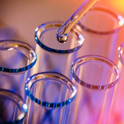 A dropper is placing droplets of chemicls in to a set of test tubes photographed against a colorful background.